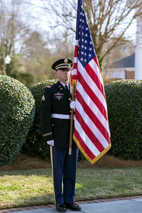 A sergeant stands at attention while holding the American flag in an outdoor area surrounded by greenery.