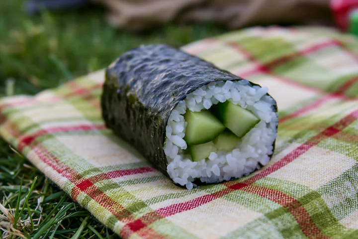 A single cucumber maki roll rests on a plaid picnic blanket, showcasing fresh cucumber pieces surrounded by sushi rice and seaweed.