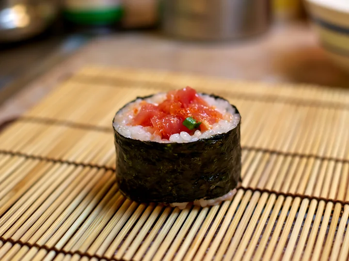 A single spicy tuna roll piece sits on a bamboo mat, showcasing the rice and tuna filling.