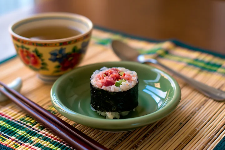 A single spicy tuna roll piece sits on a green plate next to a bowl of tea.