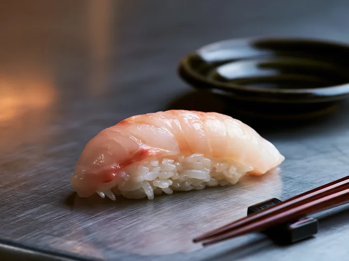 A single sushi piece sits on a modern dining table with a dark plate nearby.
