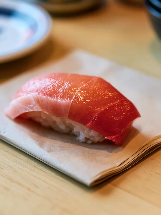 A single sushi piece with colorful fish topping rests on a light brown napkin on a wooden table.