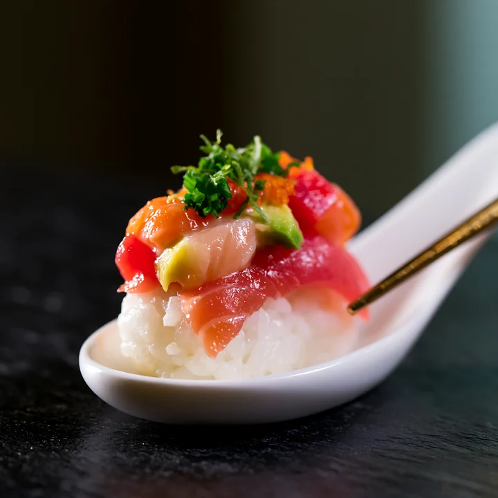 A single sushi piece with colorful fish topping rests on a white spoon.