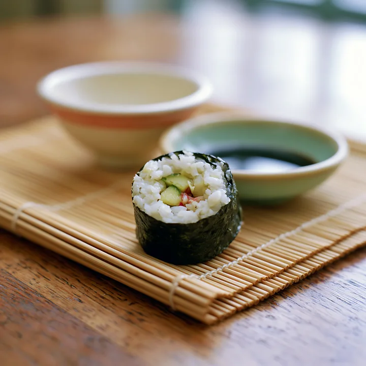 A single sushi roll on a bamboo mat is ready to eat alongside small bowls of soy sauce.