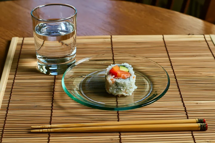 A single sushi roll on bamboo mat is placed on a clear glass plate next to a glass of water and wooden chopsticks.