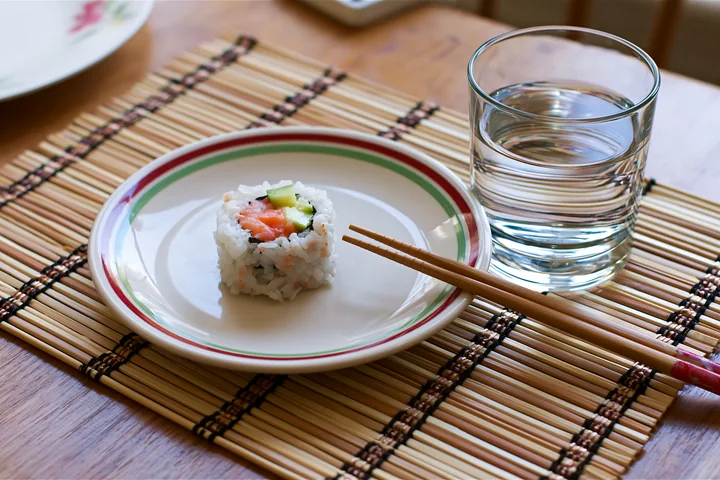 A single sushi roll on bamboo mat sits on a plate next to a glass of water and chopsticks, set on a wooden table during mealtime, showing a simple dining arrang