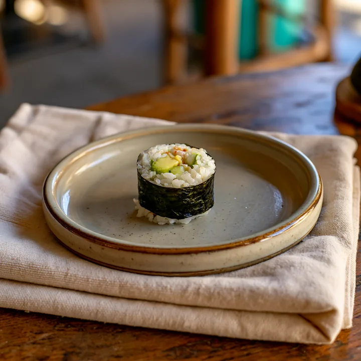 A single sushi roll on ceramic plate is placed on a white cloth napkin on a wooden table.