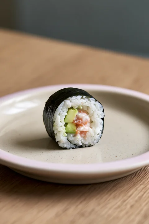 A single sushi roll on ceramic plate sits on a wooden table.