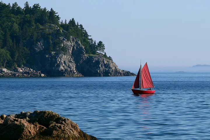 A small sailboat with red sails moves across the clear blue water near a rocky peninsula.