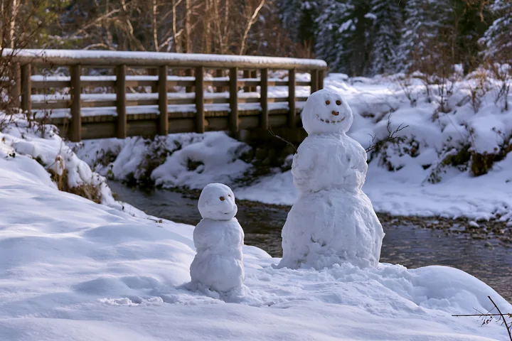 A small snowman beside a large snowman stands in the snow by a brook.