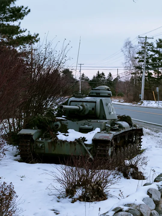 A soldier is positioned under camouflage netting near an old tank in a snowy area.