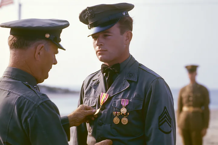 A soldier receiving a medal during an outdoor ceremony stands in uniform while a superior officer pins the medal on his chest.