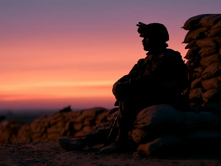 A soldier silhouette at dawn sits on sandbags, observing the horizon as the sky transitions to soft colors.