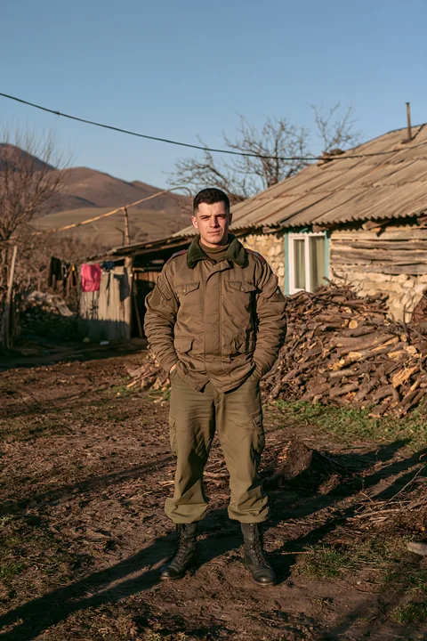 A soldier stands in front of a rustic house with a shadow on the ground.