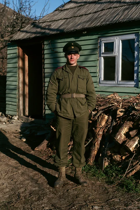 A soldier stands next to a stack of wood outside a green house.