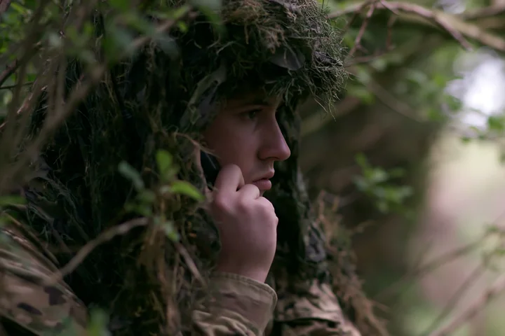 A soldier under camouflage netting speaks on the radio while hidden in dense greenery.
