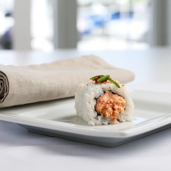 A spicy salmon sushi roll with chili sits on a white plate next to a cloth napkin.