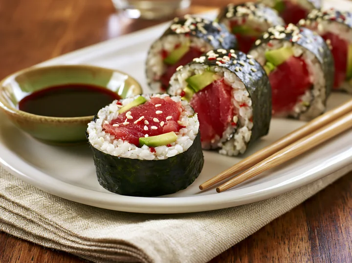 A spicy tuna roll close-up shows sushi with tuna, rice, and vegetables, placed on a white plate with soy sauce and chopsticks beside it.