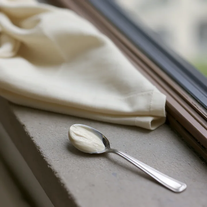 A spoon holds a small amount of cream next to a folded piece of cream-colored fabric resting on a windowsill.