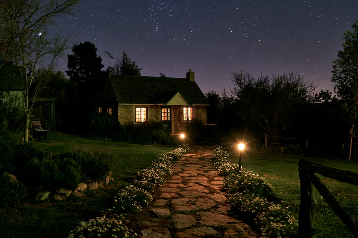 A stone path leads to a cottage surrounded by flowers under a starry sky.