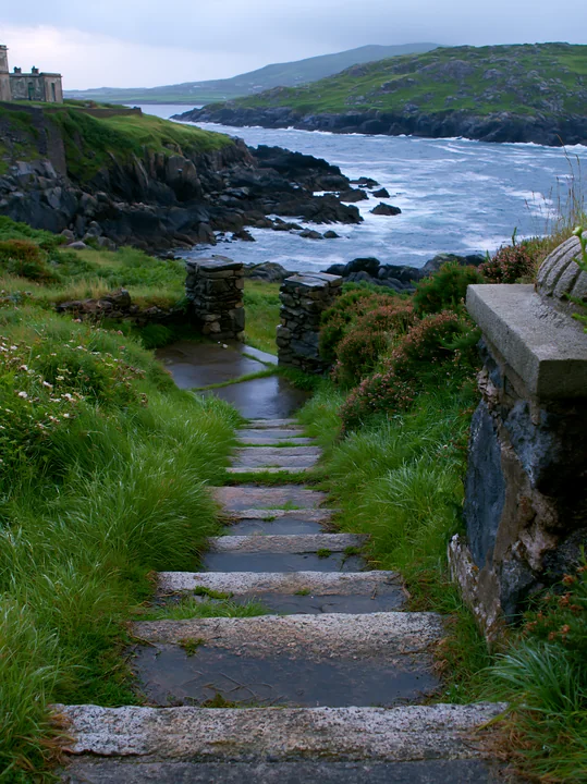 A stone pathway leads down to a rocky shoreline and an abandoned lighthouse on a remote coast.