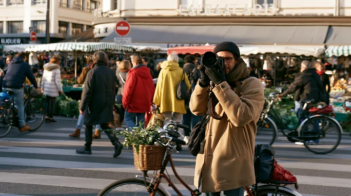 A street photographer taking pictures in an urban market setting captures the bustling scene with various people browsing stalls and bicycles in view.