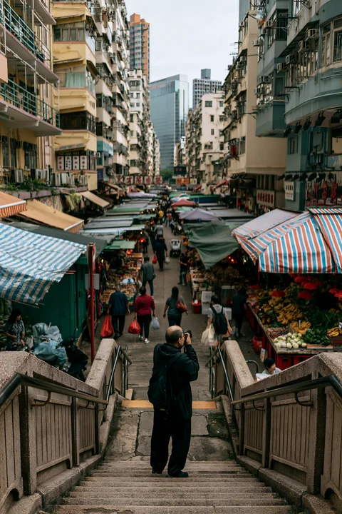 A street photographer taking pictures in an urban market stands at the top of stairs overlooking stalls filled with fresh produce and shoppers.