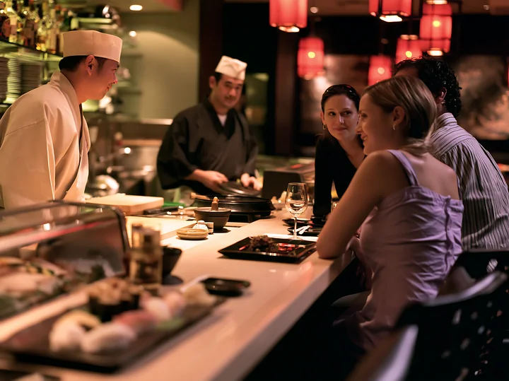 A sushi chef greeting diners at the bar while preparing food in a vibrant restaurant setting.