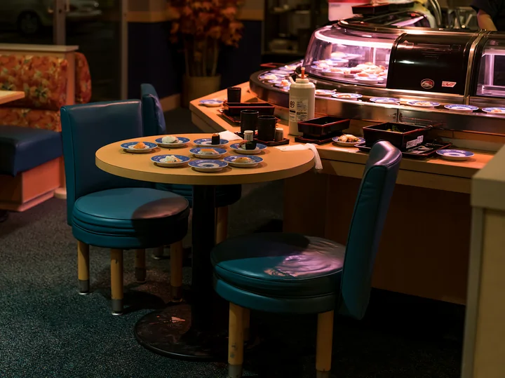 A table with plates of sushi sits near a busy sushi conveyor belt restaurant.