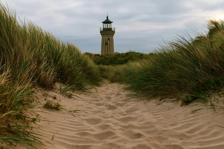 A tall lighthouse stands in the background, surrounded by coastal dunes and tall grass.