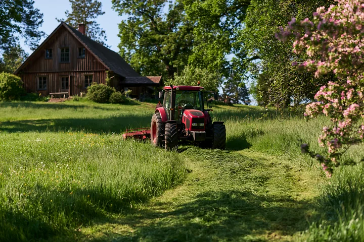 A tractor is mowing a lush green meadow next to a wooden house surrounded by trees and flowers.