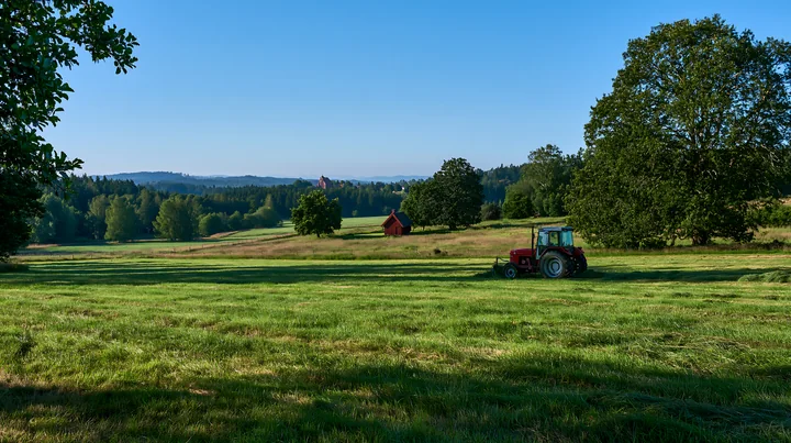 A tractor mows a green meadow in an open field surrounded by trees.