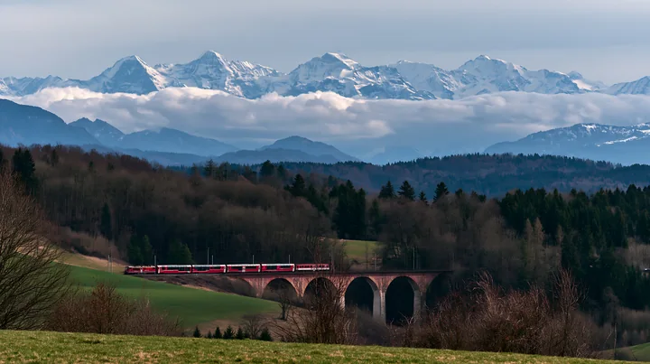 A train crosses a stone viaduct with dramatic clouds around alpine peaks in the background.
