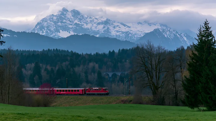 A train travels along a track with dramatic clouds around alpine peaks in the background.