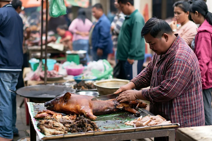 A vendor prepares a spit-roasted suckling pig with crackling skin at a busy market stall.
