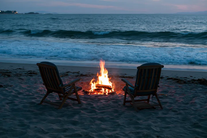 A warm fire burns in a circle of stones on the beach, with two chairs positioned nearby.