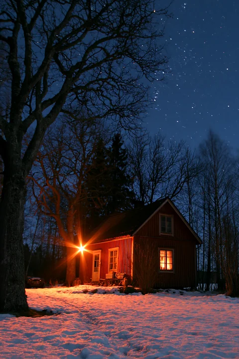A warm light glows from the windows of a small home surrounded by trees on a clear winter night.