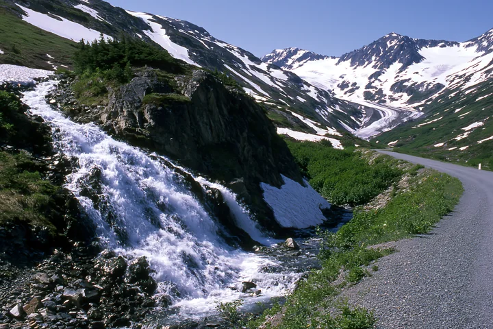 A waterfall cascades over rocky terrain with snow-capped mountains in the background.