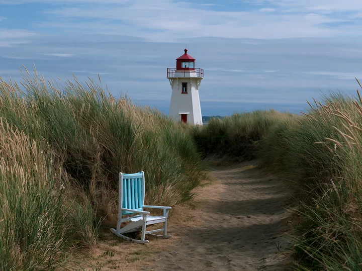A white lighthouse stands tall amid coastal dunes with tall grass, while a blue chair is positioned along a sandy path leading towards the structure.