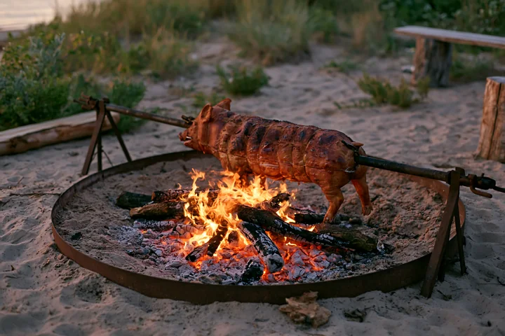 A whole pig is being roasted on a spit over an open fire in a sandy area near a body of water.