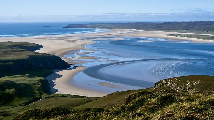 A wide sandy beach bordered by water, set against rolling hills under a clear blue sky.