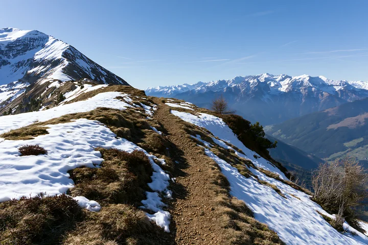 A winding trail stretches along a high mountain ridge with snow patches.