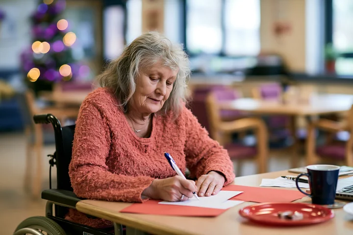 A woman in a wheelchair is writing a card at a table in a community center.