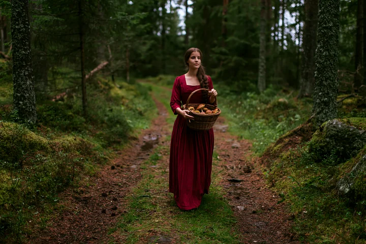 A woman in traditional dress walks along a forest path, carrying a basket of mushrooms.
