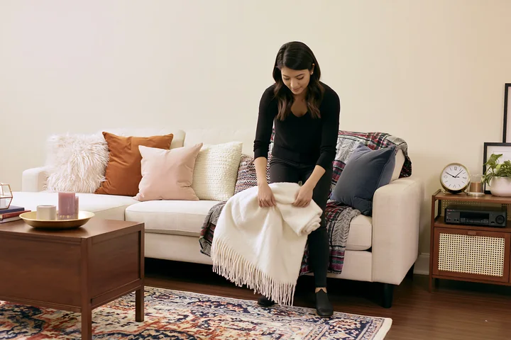 A woman is folding a white blanket on a sofa in a cozy living room.
