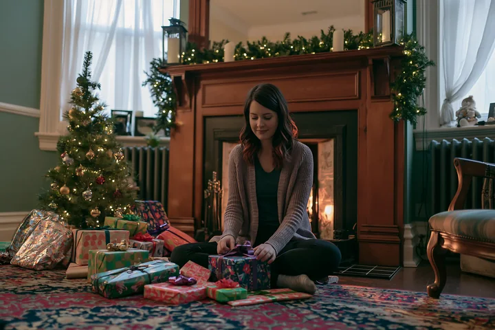 A woman sits on the floor in a living room surrounded by wrapped gifts and a small Christmas tree, with a wooden mantel and warm fire in the background.
