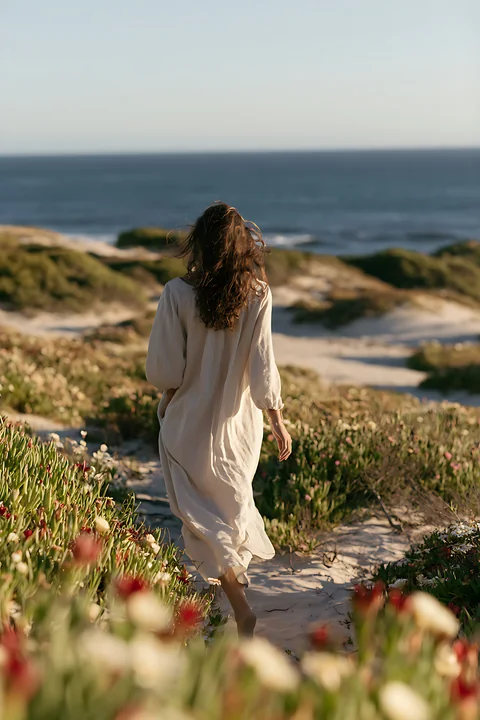 A woman walks along a sandy path bordered by flowers, heading towards the blank horizon of the ocean under a clear sky.