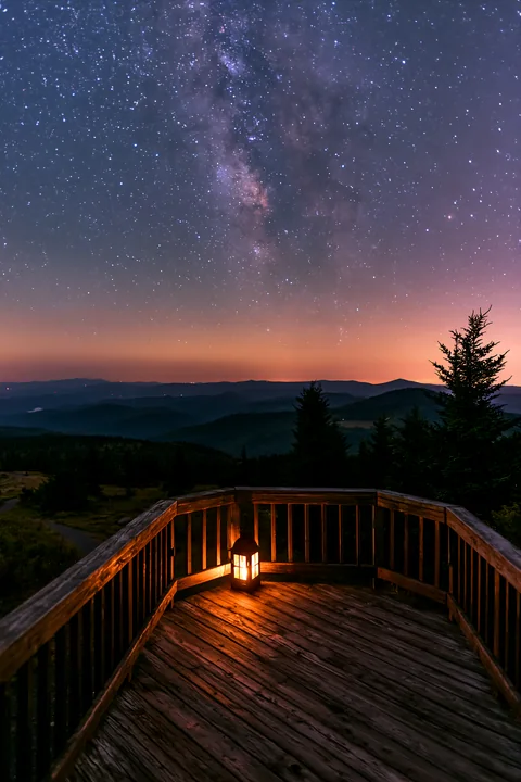 A wooden deck overlooks a mountain valley under a starry night sky.
