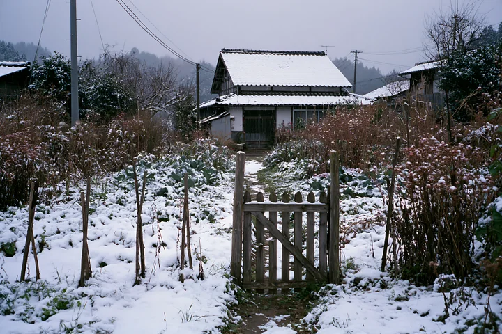 A wooden gate opens to a snowy pathway leading to a country house surrounded by a winter landscape.
