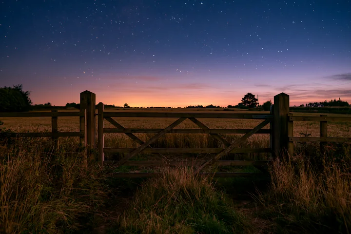 A wooden gate stands open at the edge of a wheat field under a starry night sky.